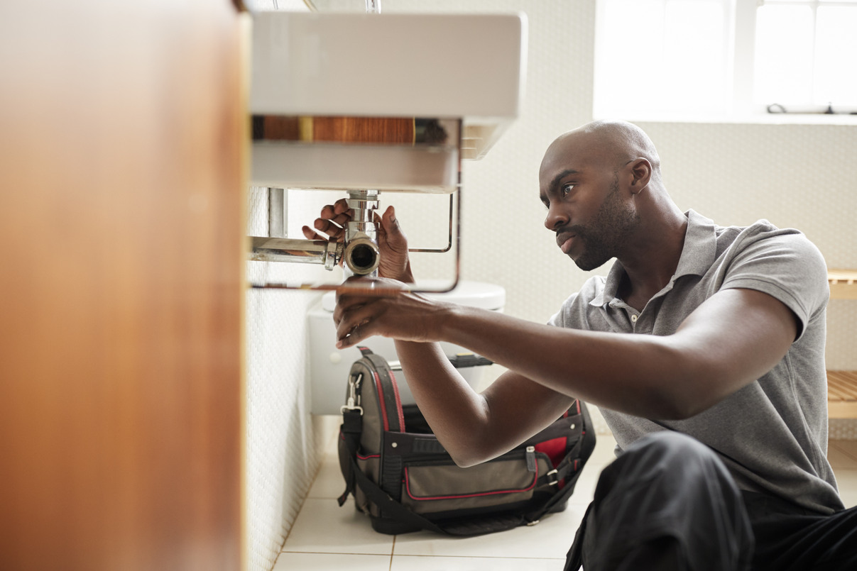 A man wearing a gray polo shirt fixing a bathroom sink’s plumbing with a wrench while sitting on the floor, with a tool bag nearby.