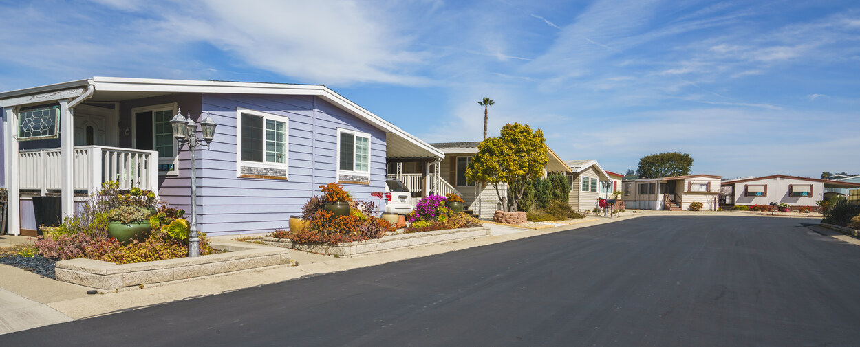 Row of manufactured homes in a mobile home park with clean asphalt road, landscaped yards, and clear blue sky.