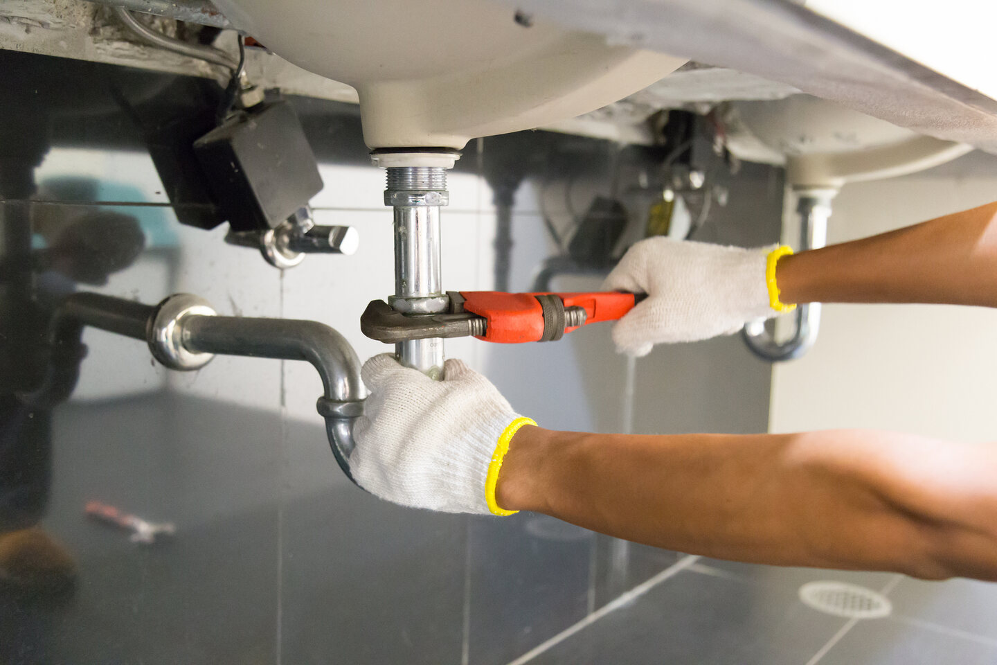 Close-up of a person wearing white gloves using a red pipe wrench to tighten or loosen a pipe under a sink.