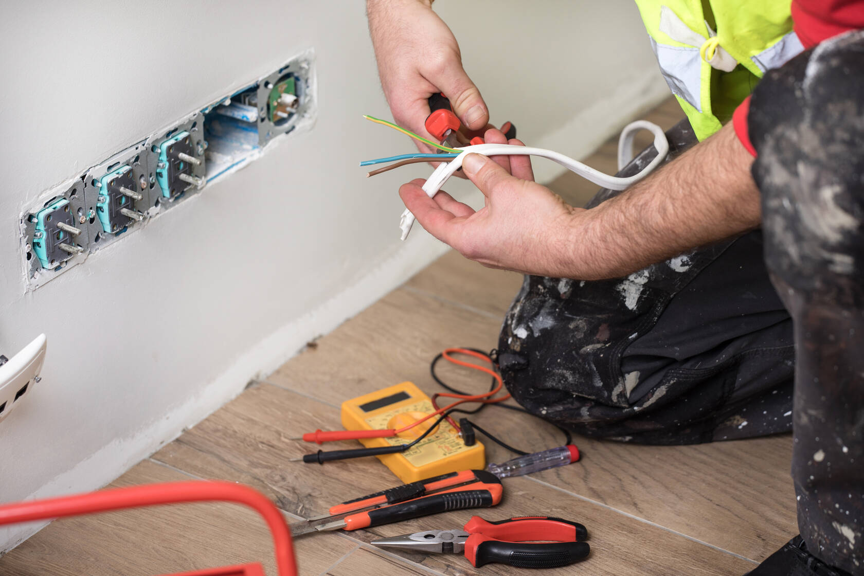 Electrician kneeling on a wooden floor using pliers to strip wires from a white cable near an unfinished wall outlet with exposed sockets and electrical tools scattered on the floor.