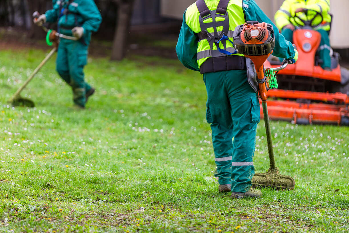 Three landscapers in green uniforms and yellow safety vests working on a grassy area, using string trimmers and a ride-on mower for lawn maintenance.