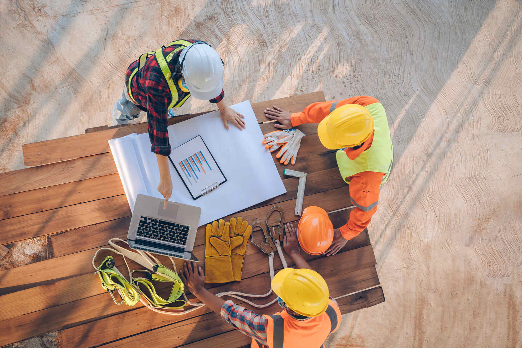 Three construction workers wearing hard hats and safety vests gathered around a wooden table with blueprints, a laptop, protective gloves, and safety harnesses, reviewing plans at a construction site.