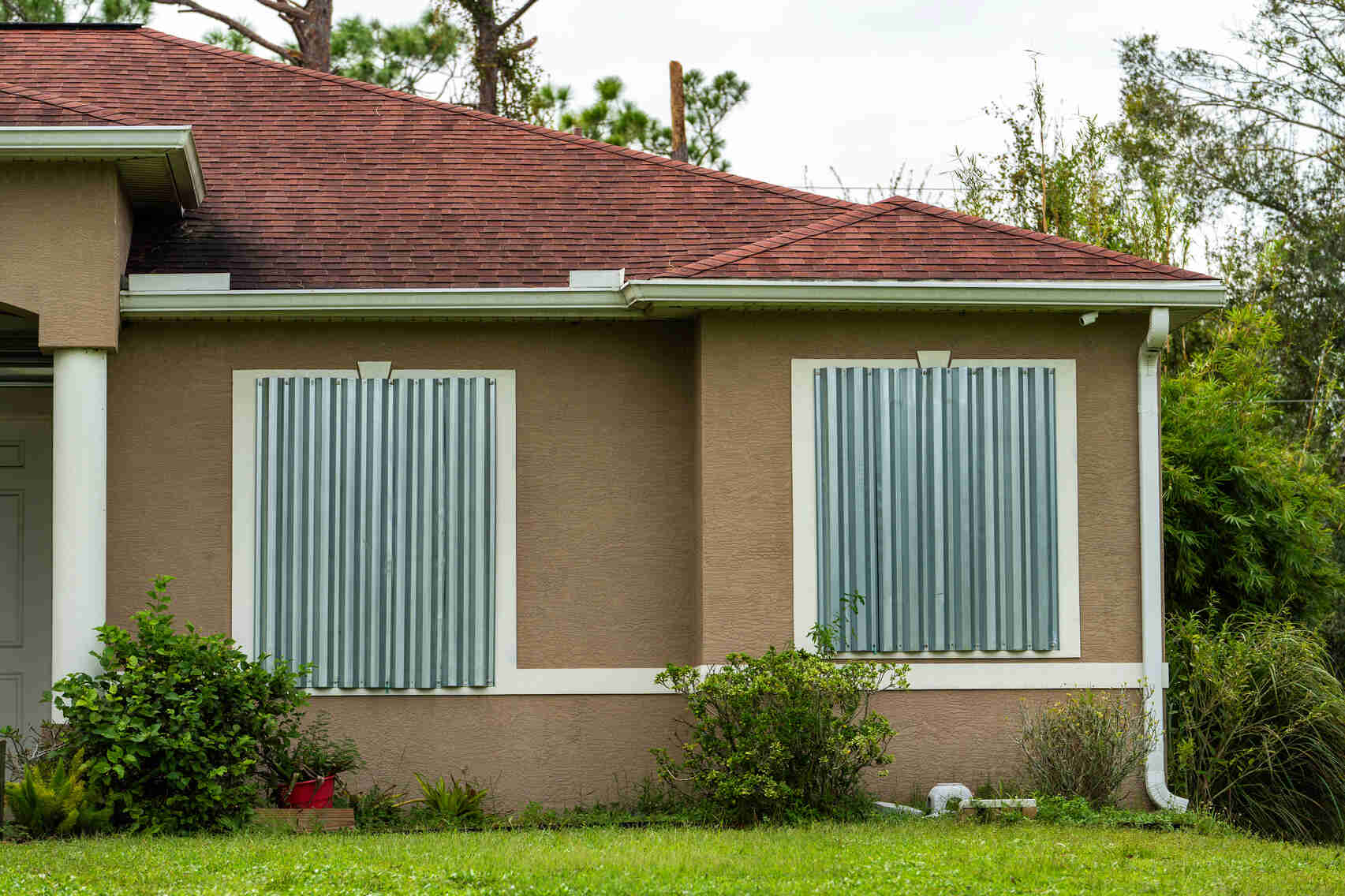 House with two windows boarded up by metal storm shutters, surrounded by green shrubbery and lawn under an overcast sky.