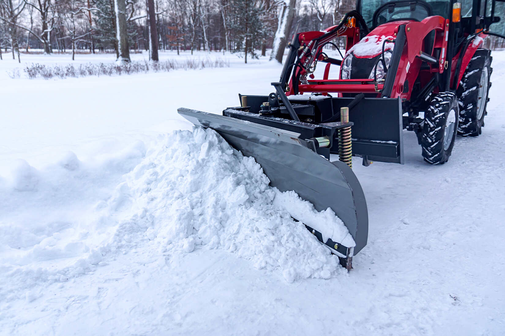 Red tractor with a front-mounted snow plow clearing snow on a snowy path in a winter landscape with trees in the background.