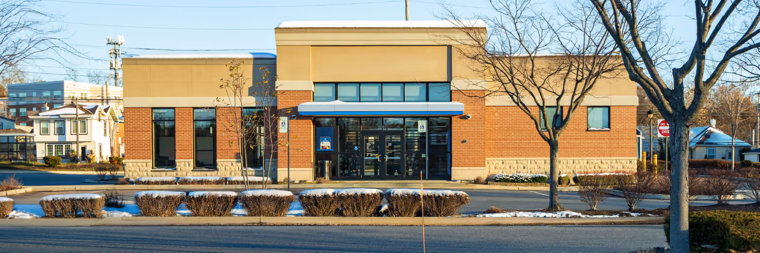 Single-story commercial building with brick and beige exterior, glass entrance with blue awning, surrounded by leafless trees and shrubs, under a clear sky.