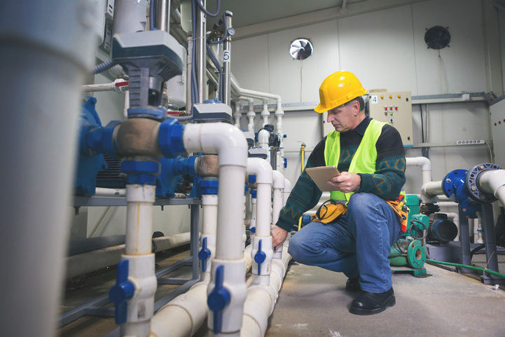 Plumber wearing a yellow hard hat and reflective vest crouches inspecting white plumbing pipes with blue valves in an industrial setting, holding a tablet in one hand.