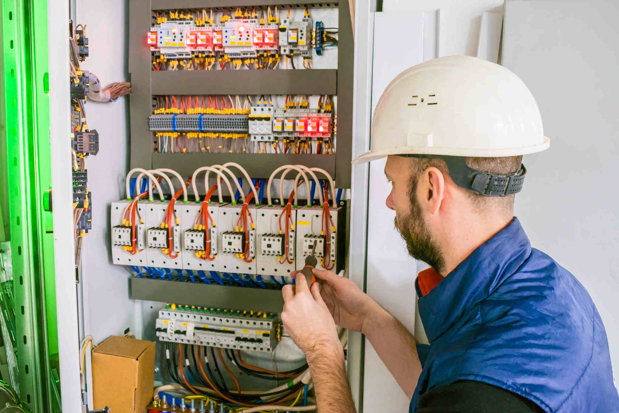 Electrician wearing a white hard hat and blue vest working on a complex electrical panel with multiple wires and circuit breakers, using a screwdriver.