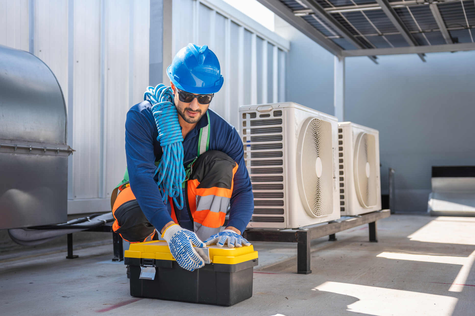 A male HVAC technician wearing a blue hard hat, sunglasses, and work gloves opens a yellow and black toolbox outside near two large air conditioning units while carrying a blue coiled rope over his shoulder.