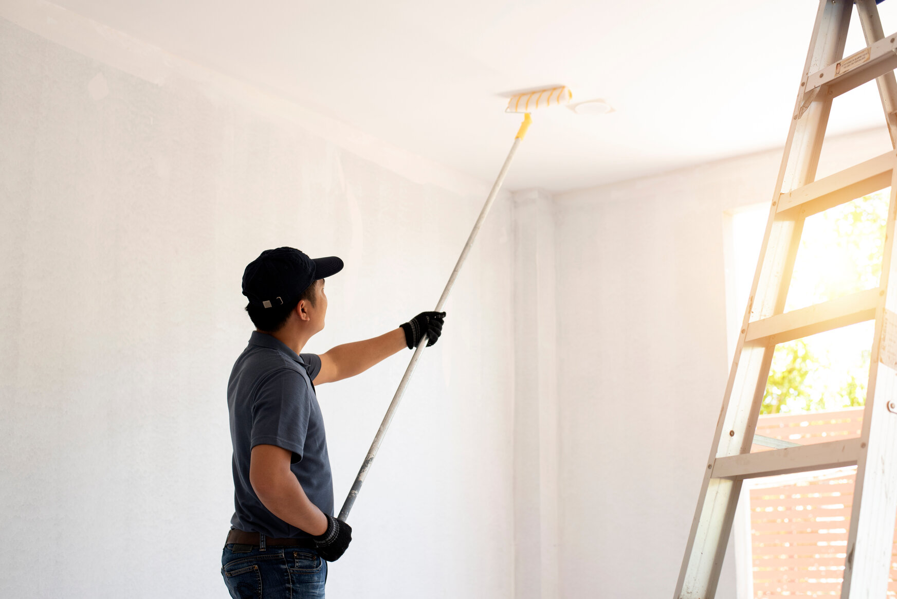 Man wearing a navy cap and gloves painting a white ceiling with a roller attached to an extension pole in a bright room next to a ladder.