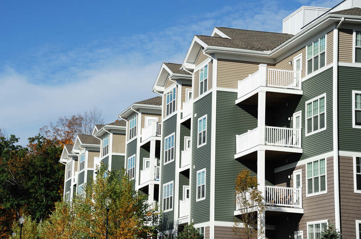 Row of modern multi-story apartment buildings with beige, green, and brown siding, white balconies, and surrounded by trees under a blue sky.