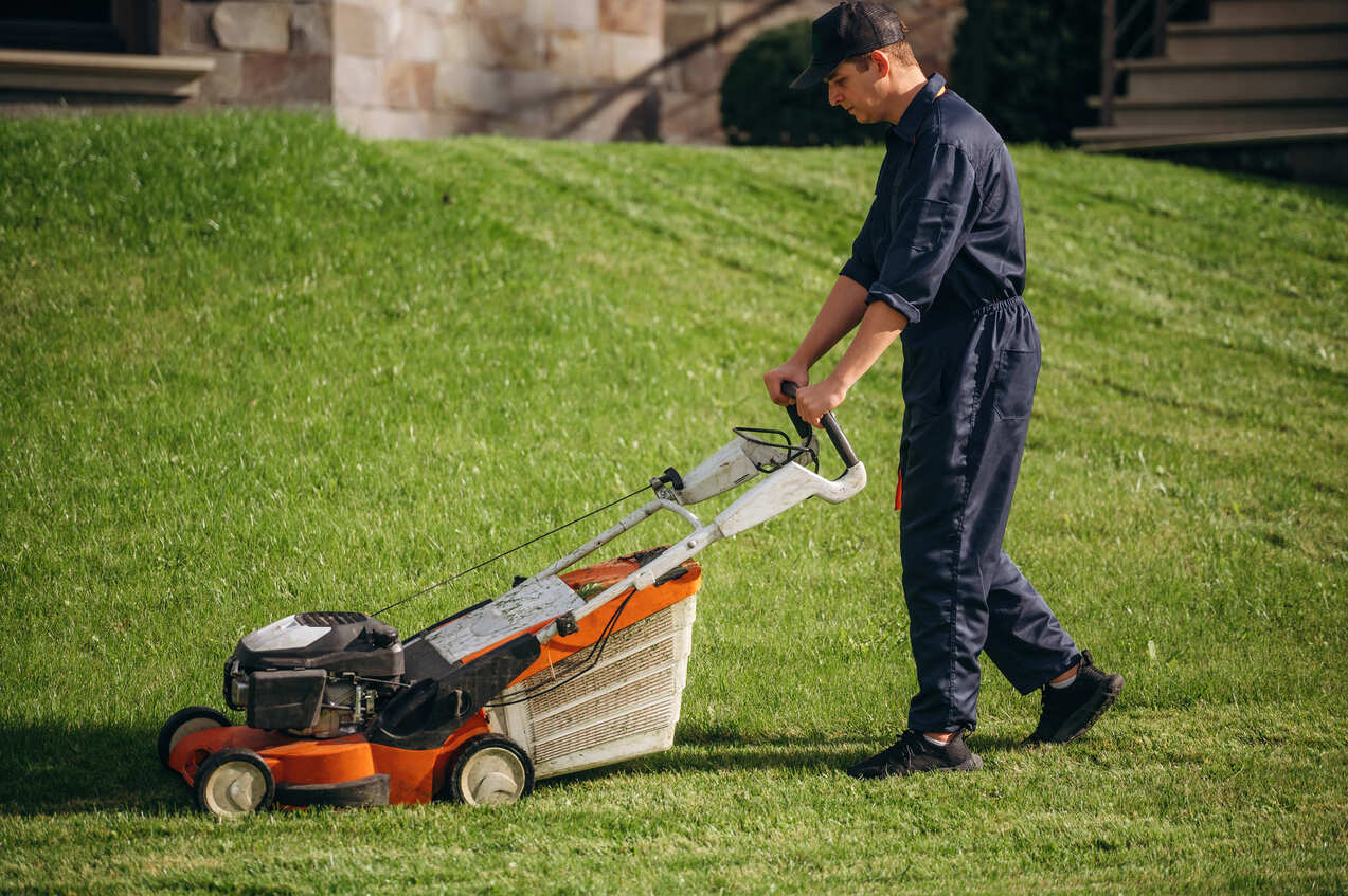 Man wearing a black cap and dark coveralls mowing a green lawn with an orange and white gas lawn mower near a stone building.