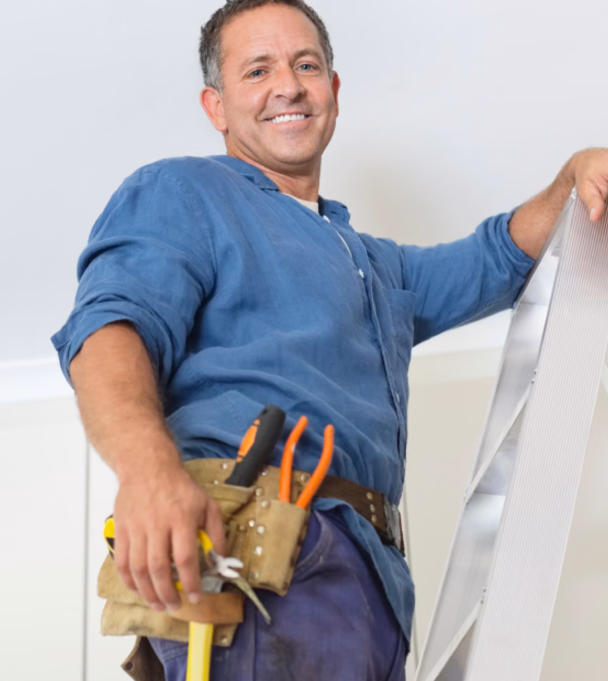 Smiling man in a blue shirt standing on a ladder with a tool belt holding pliers and a screwdriver.