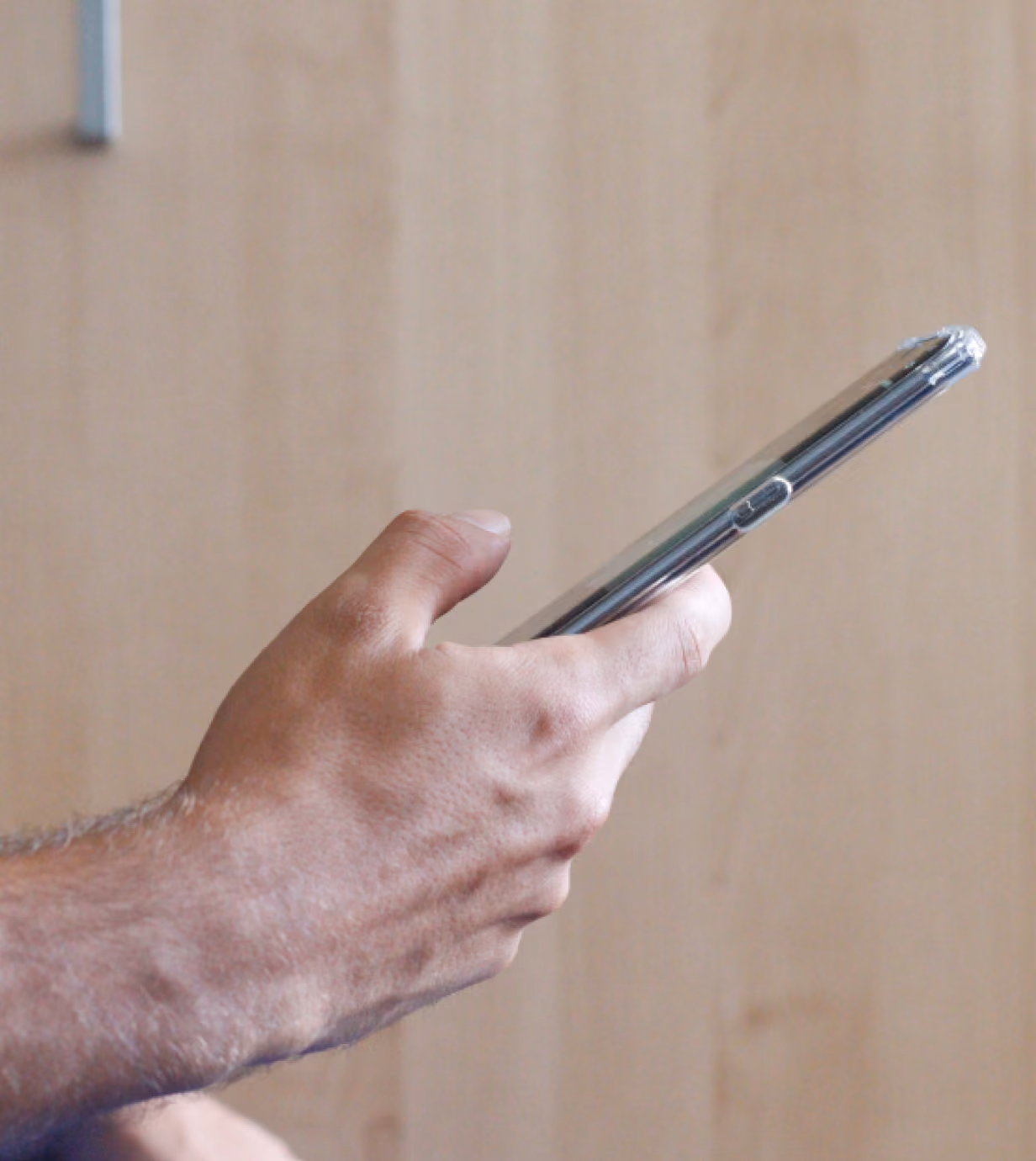 Close-up of a person's left hand holding a smartphone with a clear protective case against a plain light wood background.