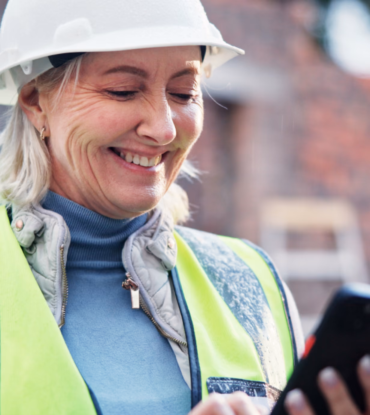 Smiling female construction worker wearing a white hard hat and neon yellow safety vest looking at a smartphone.