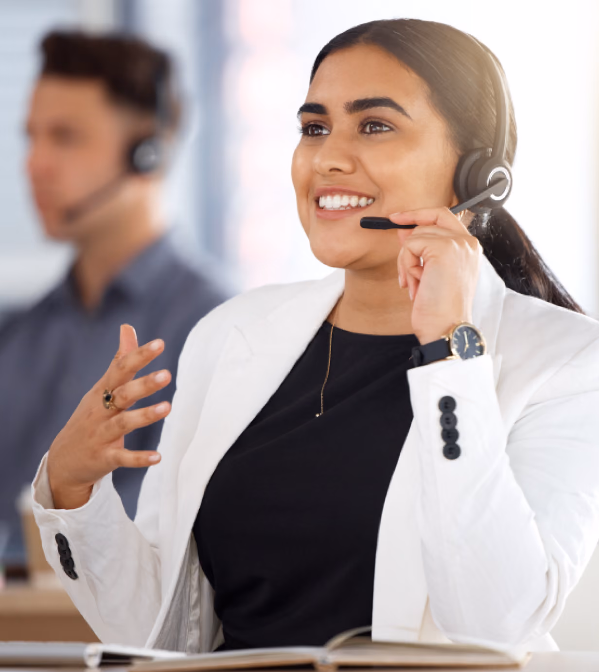 Smiling woman wearing a white blazer and a headset, speaking with hand gestures in a bright office environment with a blurred man in the background also wearing a headset.