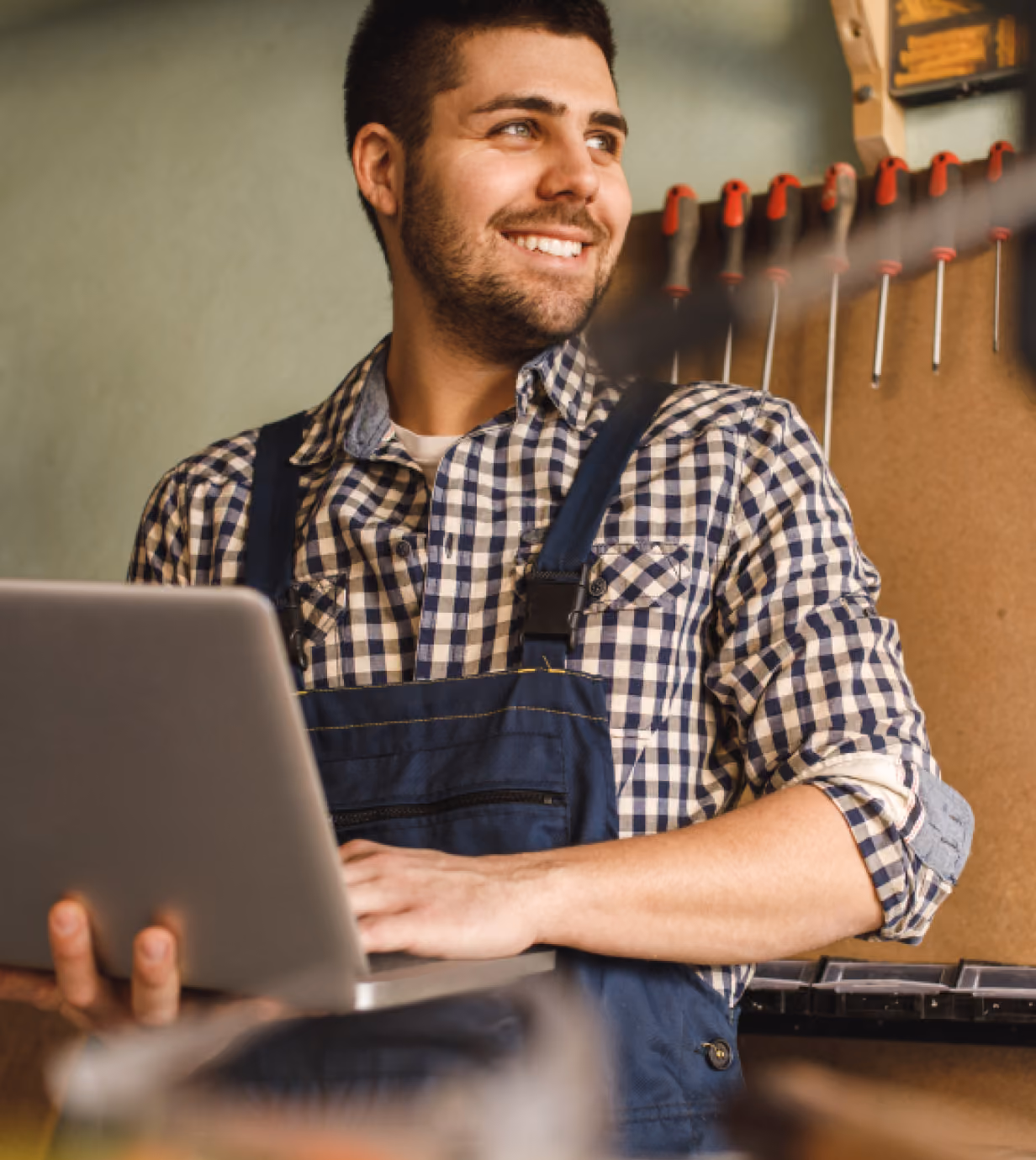 Smiling man wearing blue overalls and a checkered shirt holding a laptop in a workshop with tools hanging on the wall behind him.