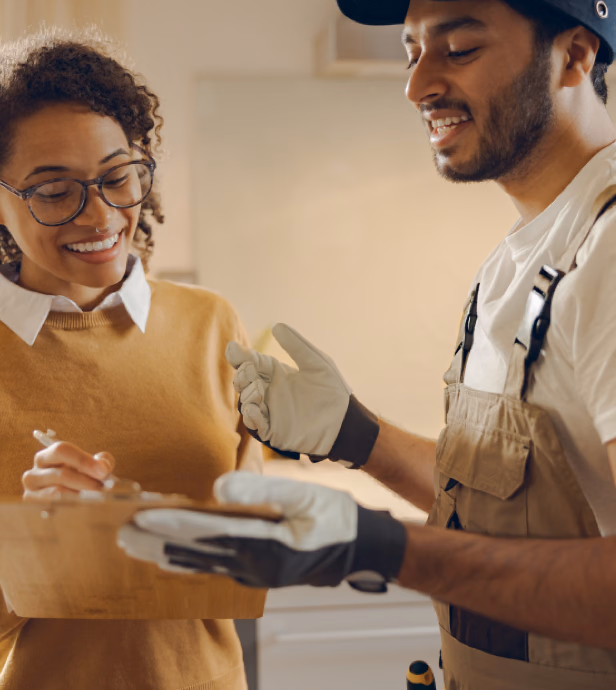 A woman smiling and holding a clipboard as a worker in overalls and gloves explains something to her indoors.
