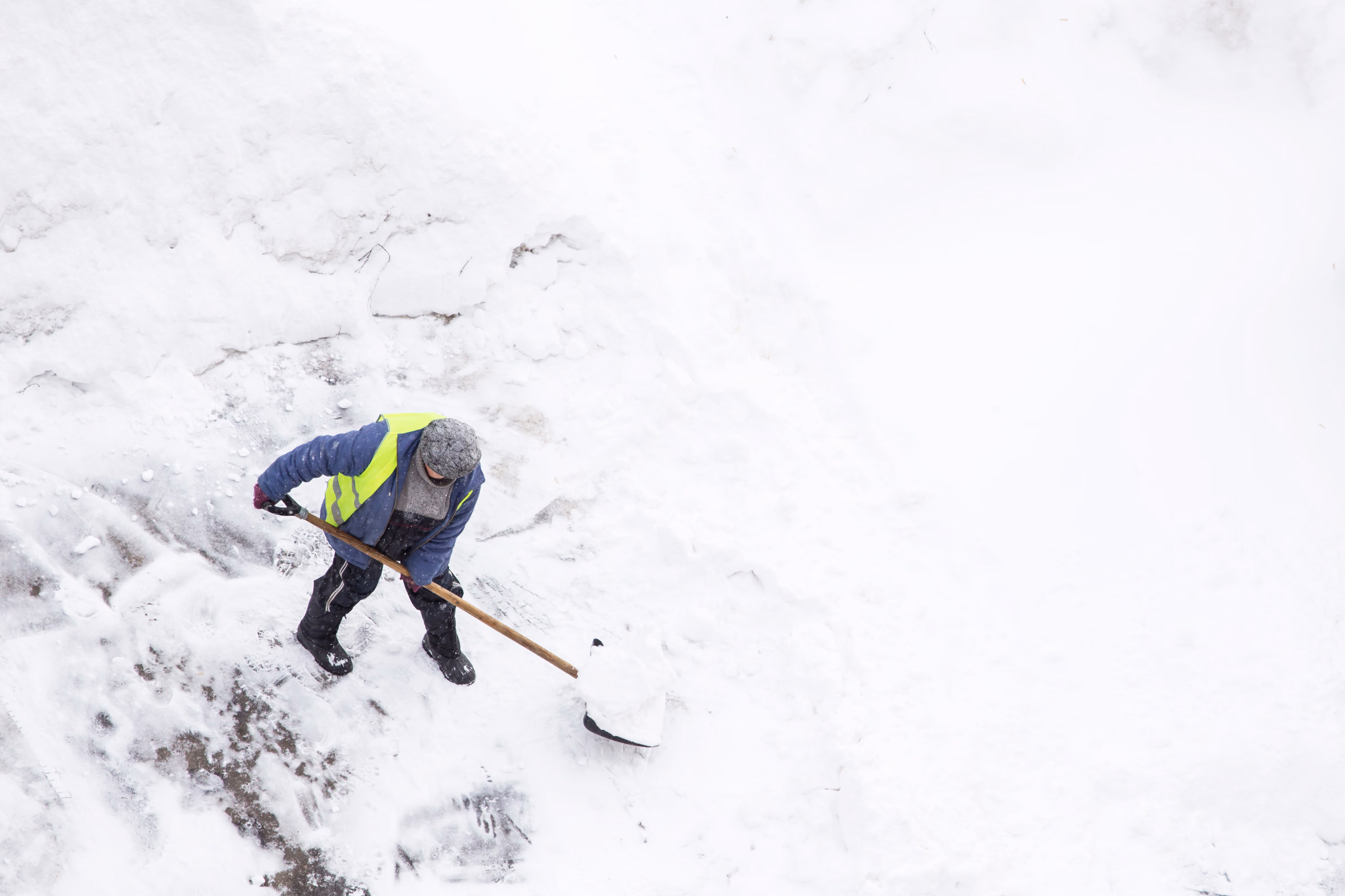 Person wearing a blue jacket and yellow reflective vest shoveling snow on a ground mostly covered in snow, viewed from above.
