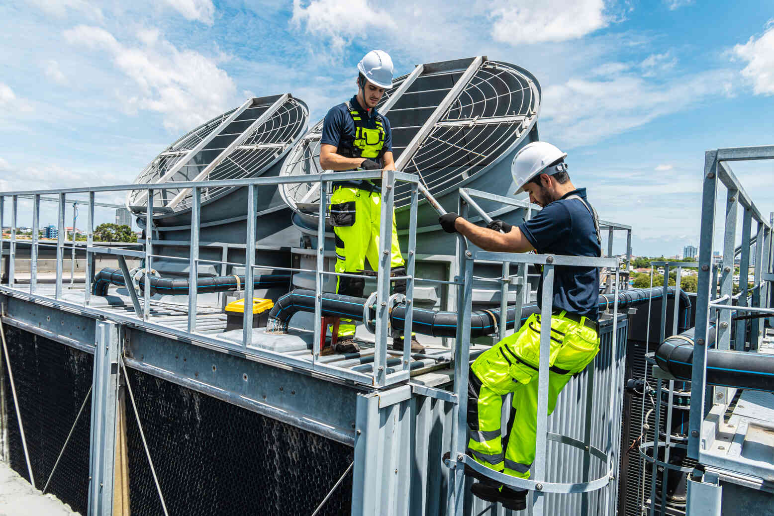 Two HVAC technicians in high-visibility yellow pants and white helmets working on rooftop ventilation equipment under a blue sky with scattered clouds.