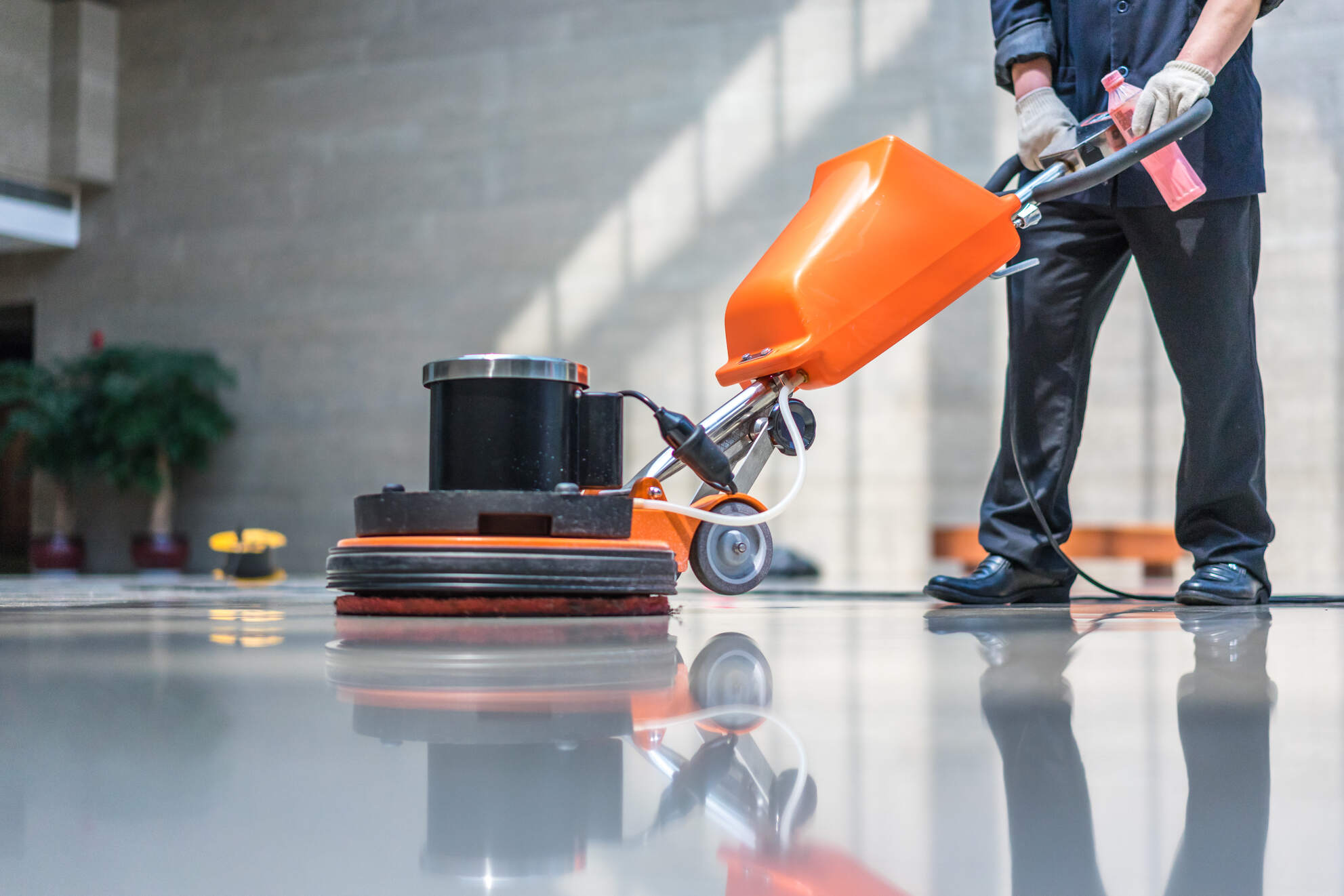 Person wearing gloves operating an orange floor polishing machine on a shiny, reflective floor in an indoor setting.