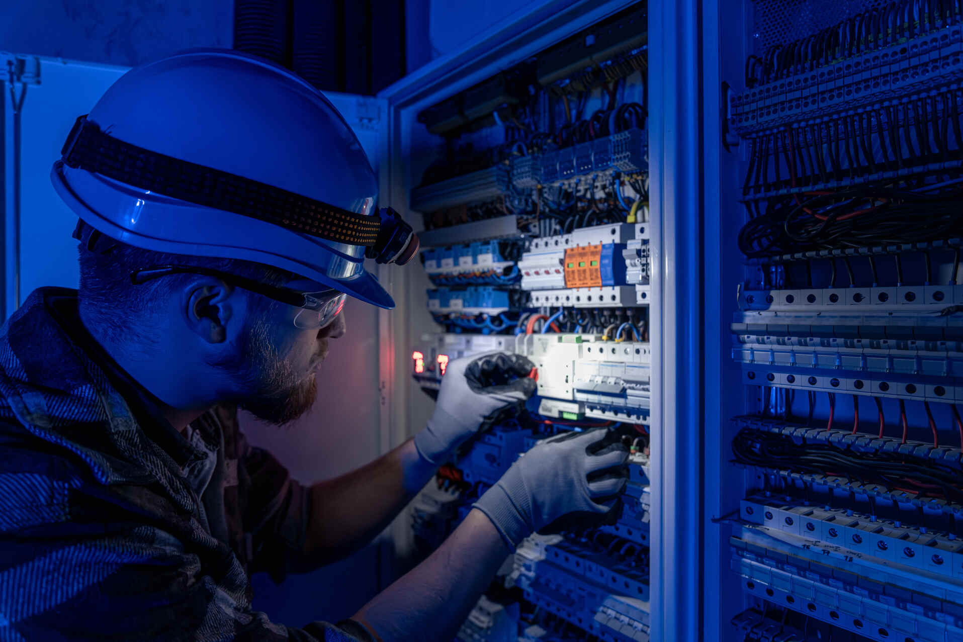 Electrician wearing a helmet with a headlamp, safety glasses, and gloves, working on wiring inside an electrical panel illuminated with blue light.