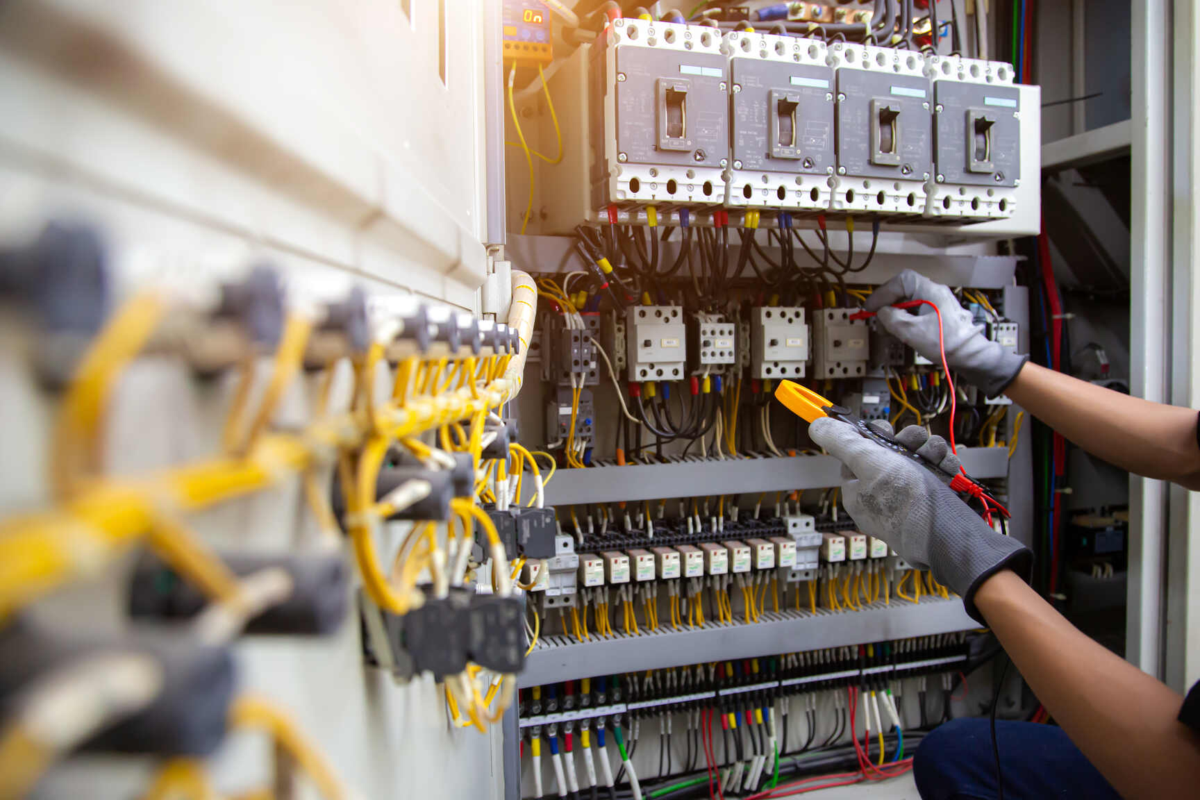 Electrician wearing gloves using a multimeter to check connections inside an electrical control panel with multiple circuit breakers and yellow wiring.