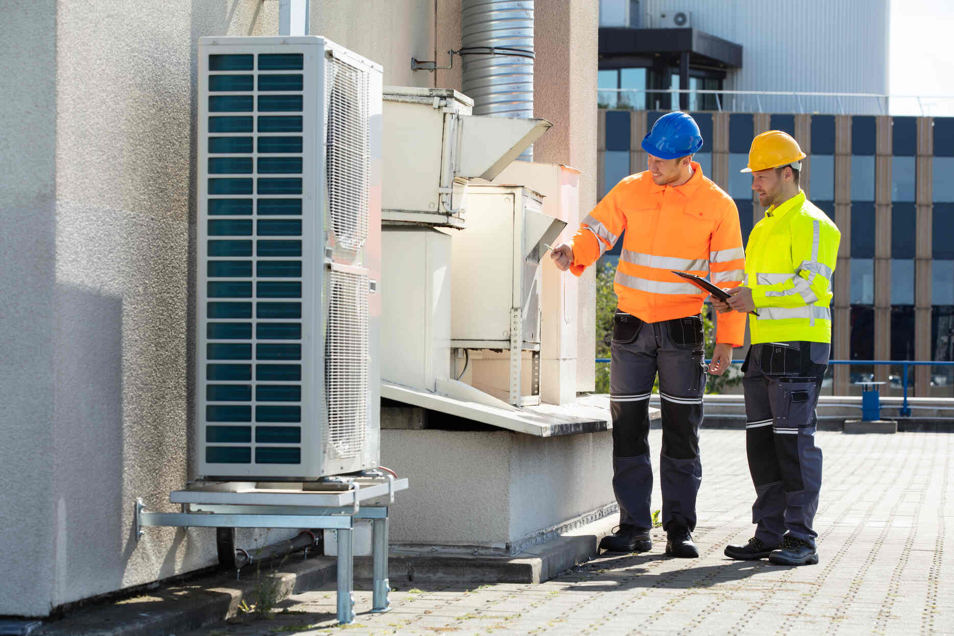 Two HVAC technicians wearing high-visibility jackets and hard hats inspecting and discussing a large air conditioning unit on a building rooftop.