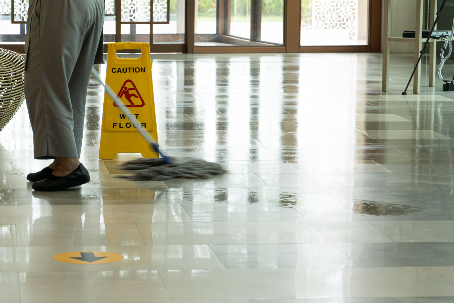 Person mopping a shiny tile floor indoors with a yellow caution sign warning of wet floor nearby.