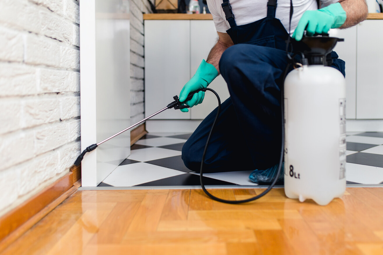 Person wearing green gloves and dark overalls kneeling on a patterned black and white tile floor while applying pest control spray along the edge of a white kitchen cabinet near a white brick wall.