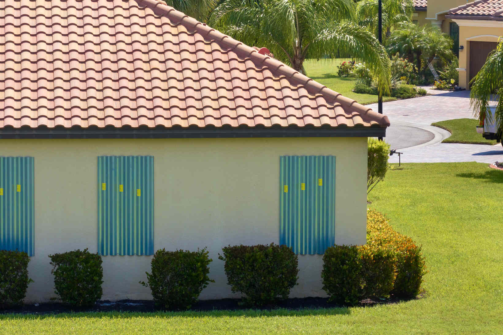 Side of a light-colored house with a red tile roof and three windows covered by blue storm shutters, surrounded by green bushes and lawn under bright sunlight.