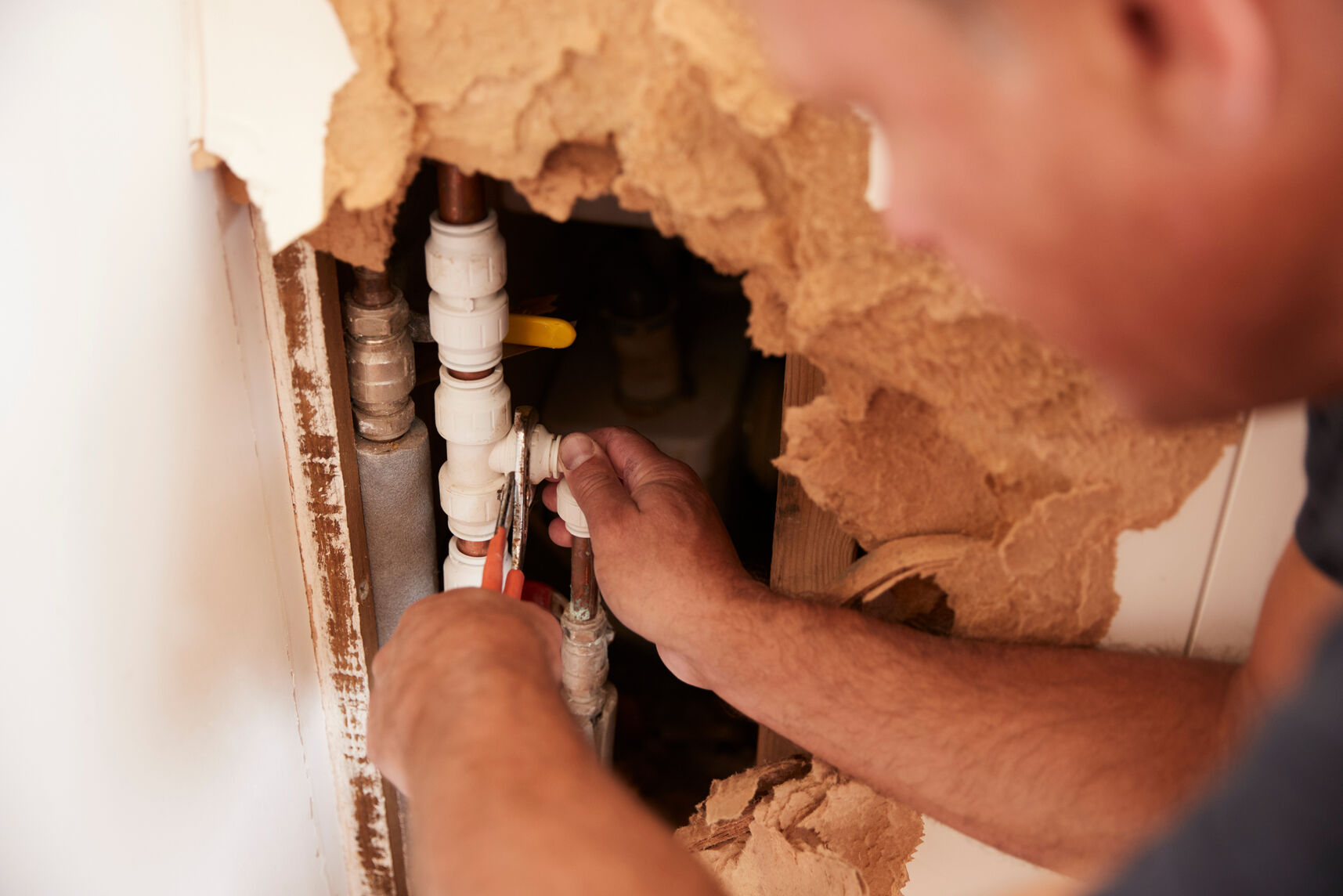 A person using a wrench to tighten or fix white plumbing pipes behind a damaged wall with torn drywall and insulation visible.