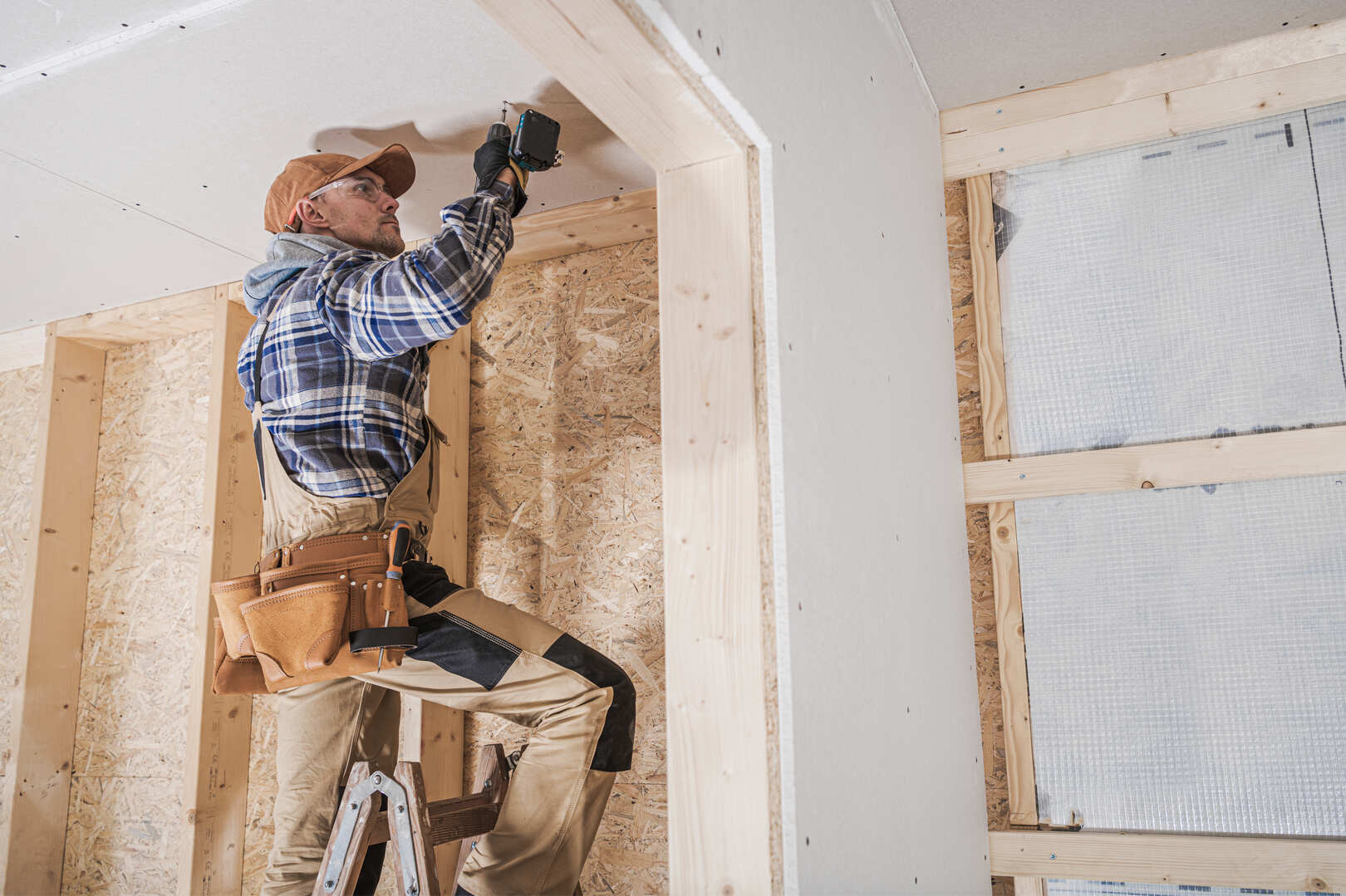 A construction worker wearing a brown cap, plaid shirt, and tool belt is standing on a ladder using a power drill to install drywall on the ceiling inside a wooden-framed room under construction.