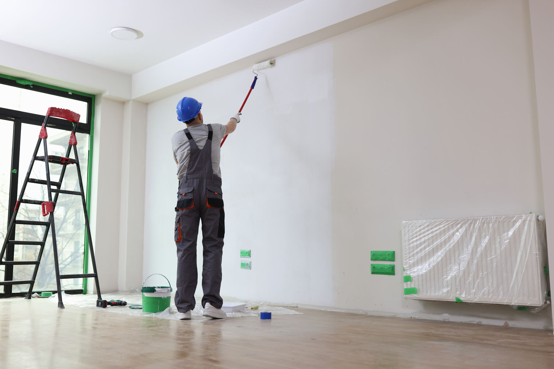 A painter in gray overalls and a blue hard hat painting a white wall with a paint roller in a room under renovation, with a ladder, paint bucket, and plastic-covered radiator visible.
