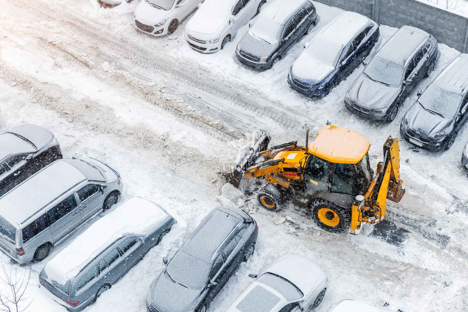 Yellow snowplow clearing snow from a parking lot filled with snow-covered parked cars during a snowfall.