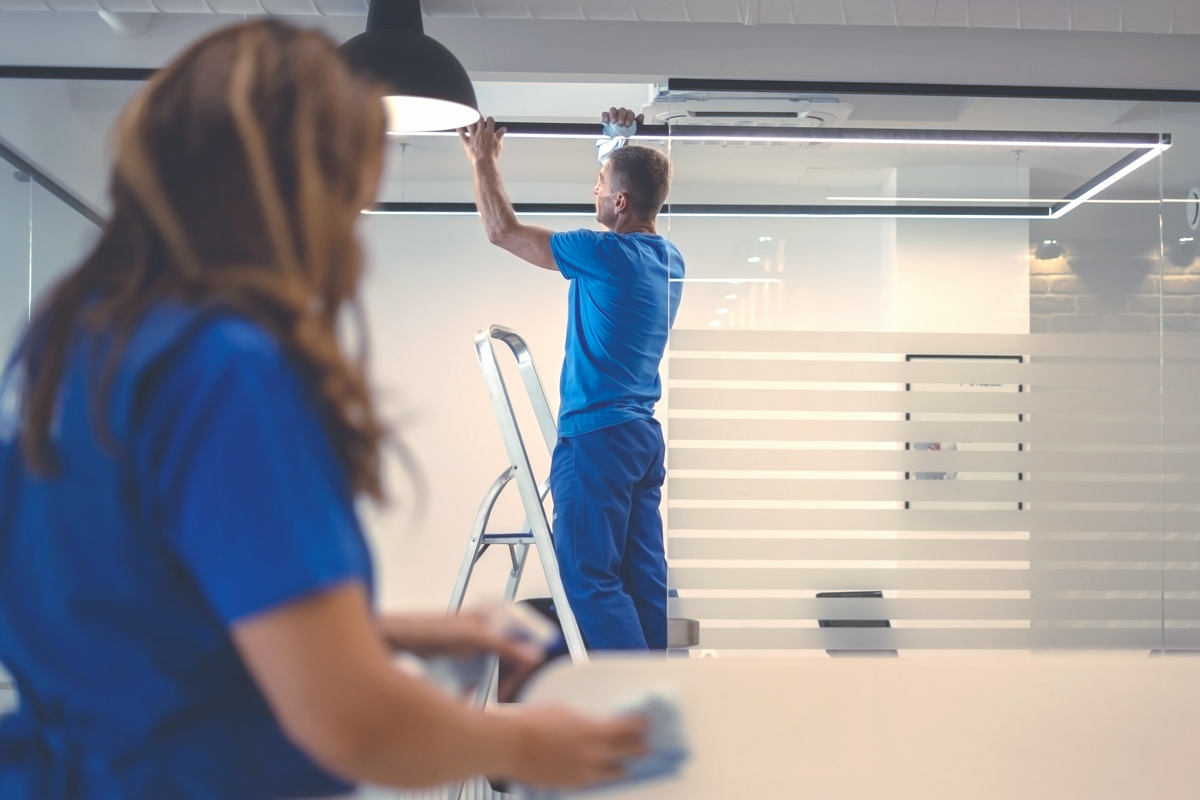 Man in blue shirt and pants standing on a ladder installing or fixing a rectangular ceiling light in a modern office, with a woman in blue scrubs cleaning a surface in the foreground.