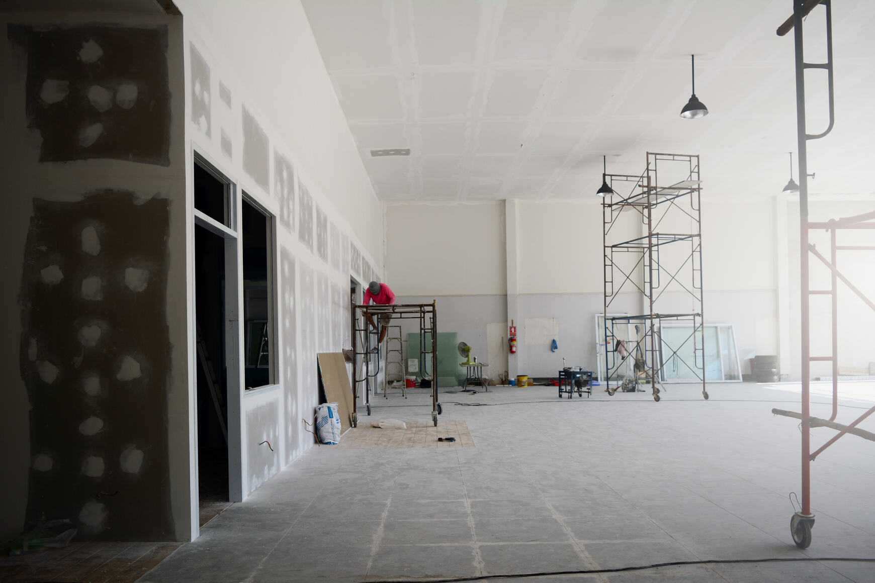 Large indoor construction site with a worker on a scaffolding near an unfinished wall, metal scaffolding structures, building materials, and ceiling lights in a spacious room with tiled floor.