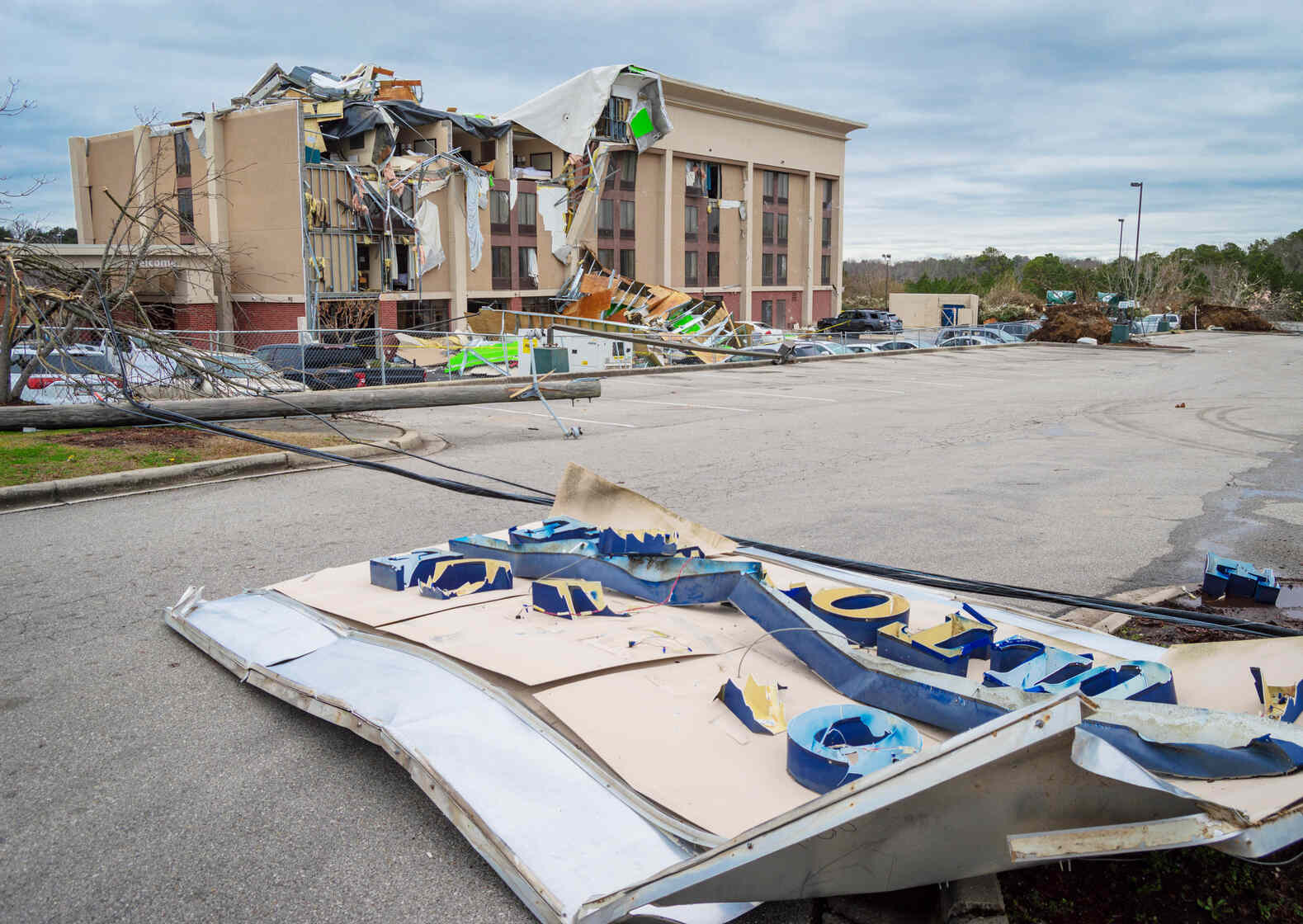 A large hotel building with extensive roof and facade damage, including exposed interior and torn materials, in the background. In the foreground, a broken and uprooted hotel sign lies on the ground. Fallen utility poles and trees with broken branches are scattered around the parking lot, indicating severe storm or tornado damage.