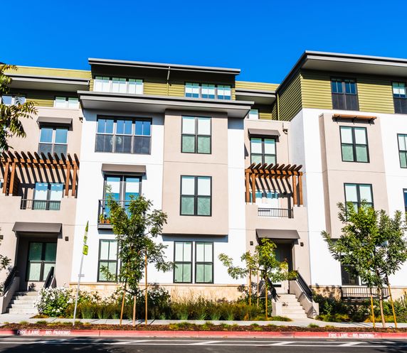 Modern multi-family residential building with white and olive green exterior, large windows, balconies with wooden pergolas, small trees, and landscaped front yard along a street with clear blue sky.