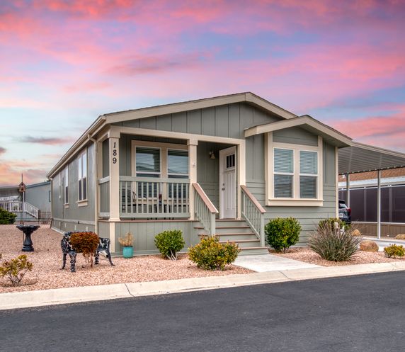 Gray manufactured home with white trim and a small porch, set against a colorful pink and purple sunset sky, surrounded by desert landscaping and gravel with decorative bushes and a metal dog sculpture.
