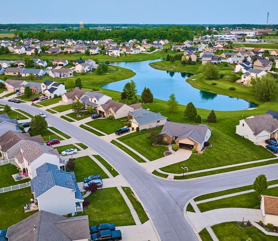 Aerial view of a suburban neighborhood with neatly maintained lawns, residential houses, winding roads, and a pond surrounded by green grass and trees.