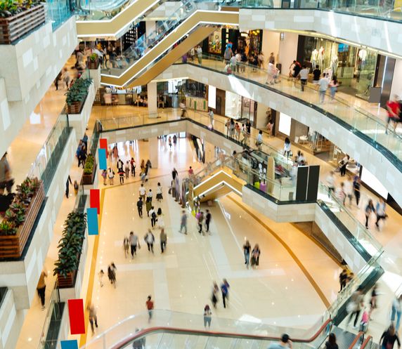 Interior view of a spacious, multi-level shopping mall with numerous people walking and shopping on bright, polished floors and glass railings.
