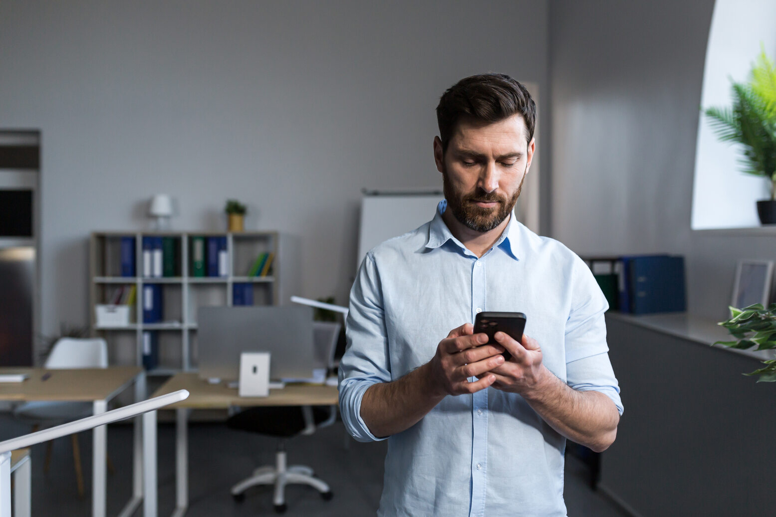 A man with dark hair and beard wearing a light blue shirt stands in a modern office looking at and using his smartphone, with desks, chairs, shelves with binders, and a plant visible in the background.
