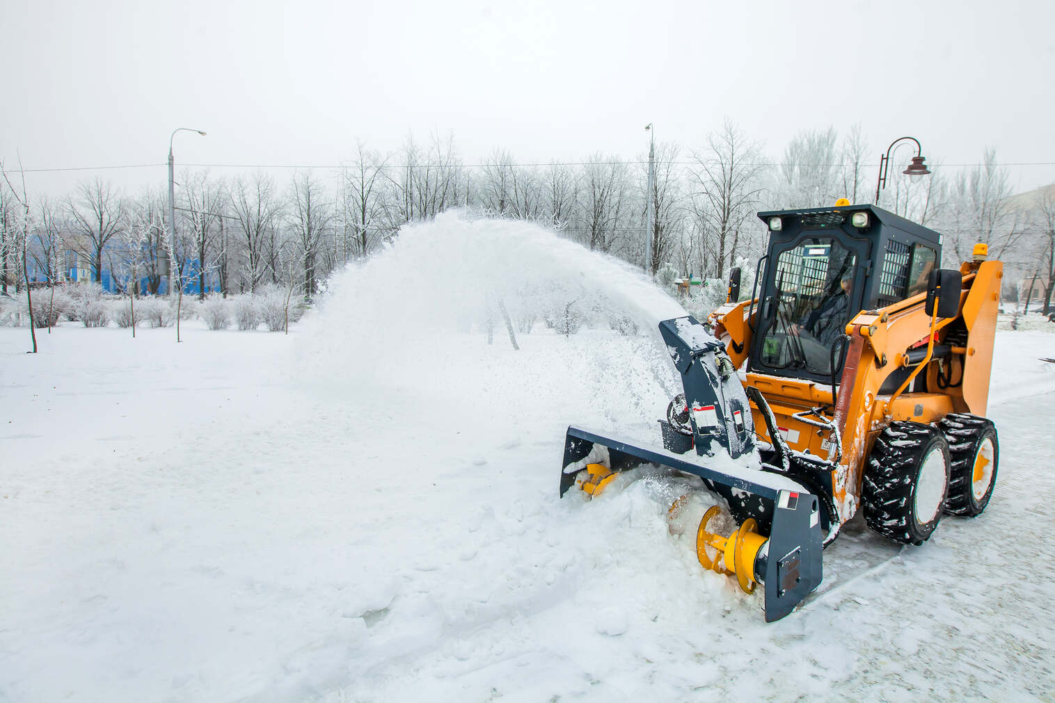 Orange snow blower vehicle clearing snow from a road in a snowy park with leafless trees and lampposts in the background.