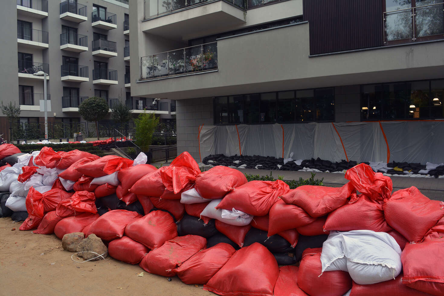 Stacked red, black, and white sandbags forming a barrier outside a modern apartment building, likely for flood protection.