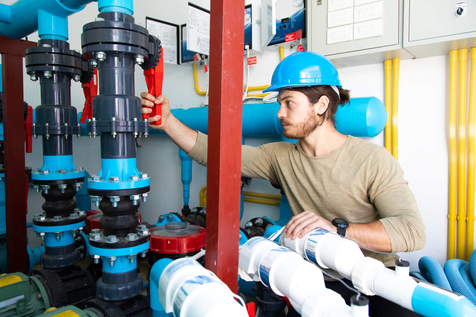 Young male technician wearing a blue hard hat and beige shirt operating a red valve on a large blue plumbing system in a mechanical room with yellow pipes and control panels.