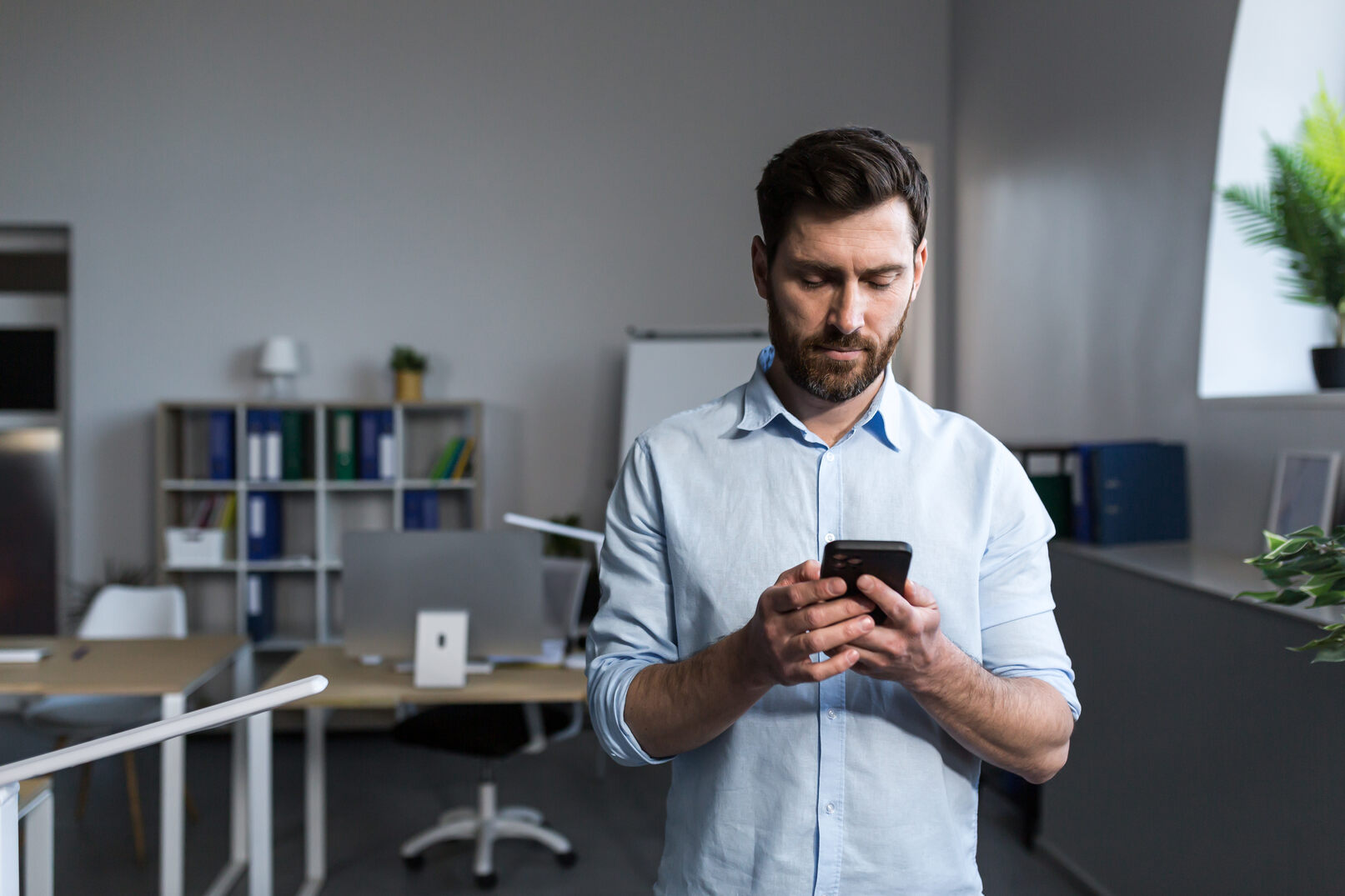 Man with beard and light blue shirt standing in an office, looking down and using a smartphone with both hands.