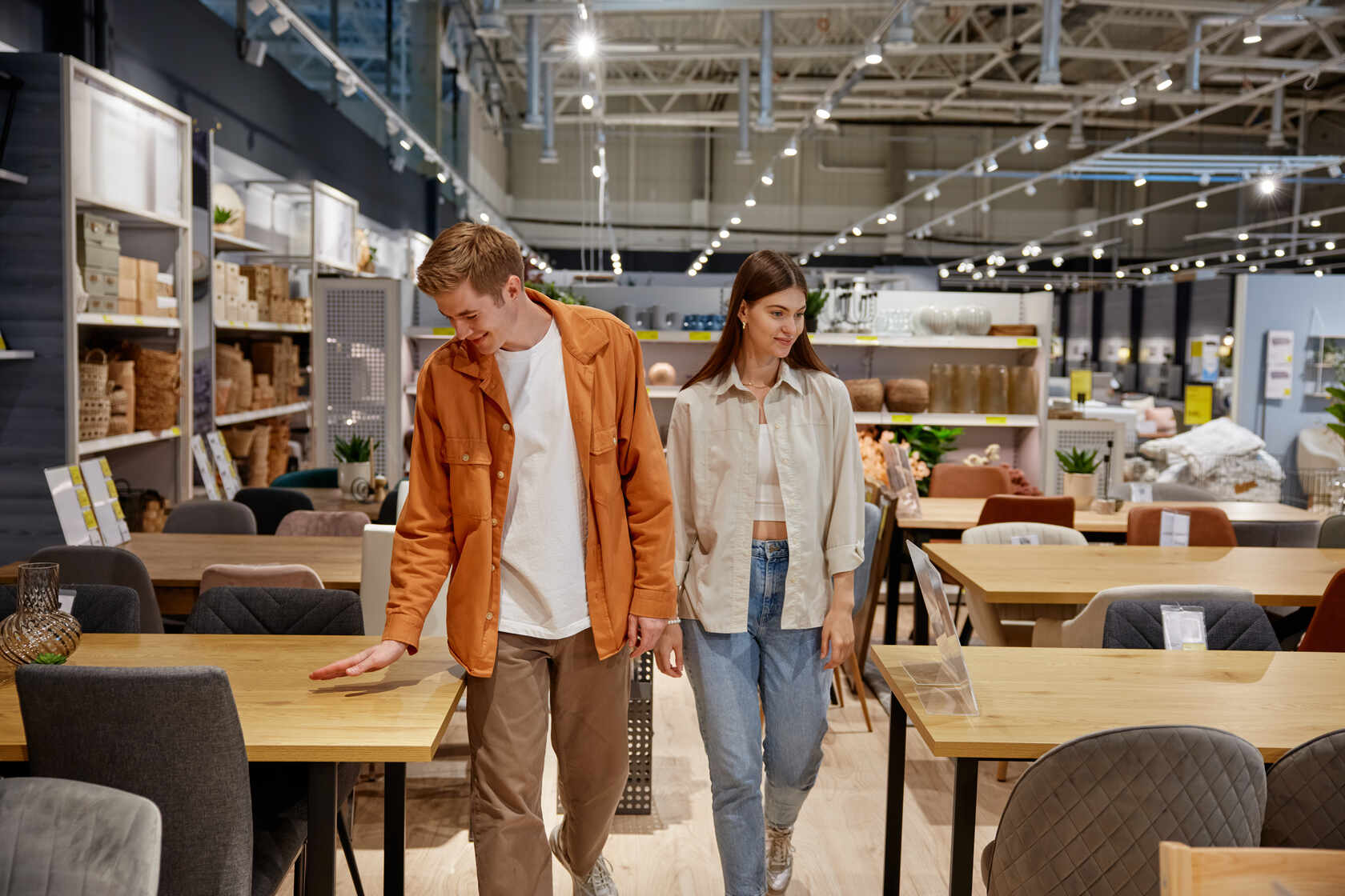 A young man and woman browsing wooden tables and chairs in a well-lit furniture store with shelves of home decor items in the background.