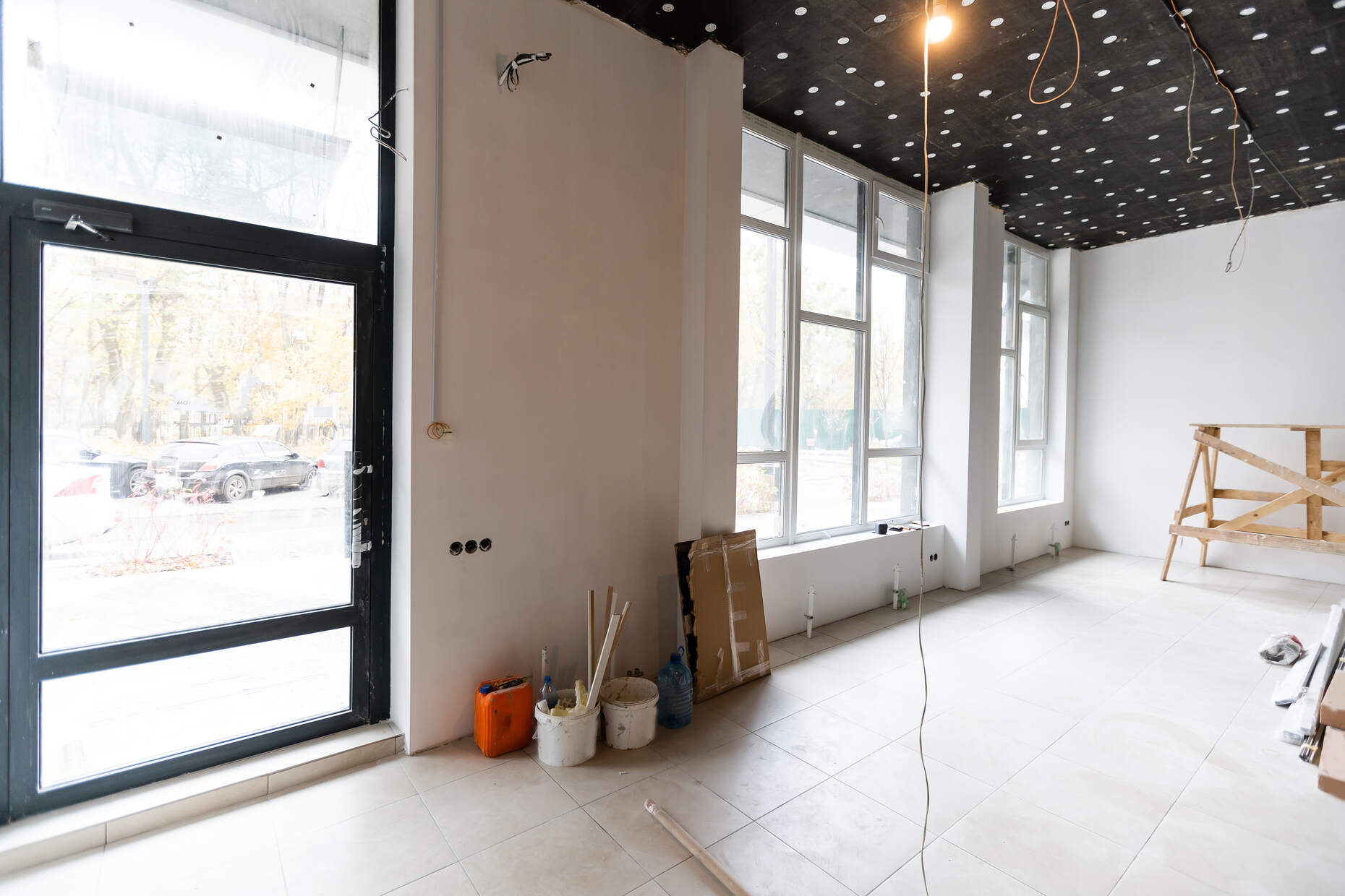 Interior of a bright room under renovation with large windows, a glass door, white tiled floor, construction materials, and exposed ceiling wiring.