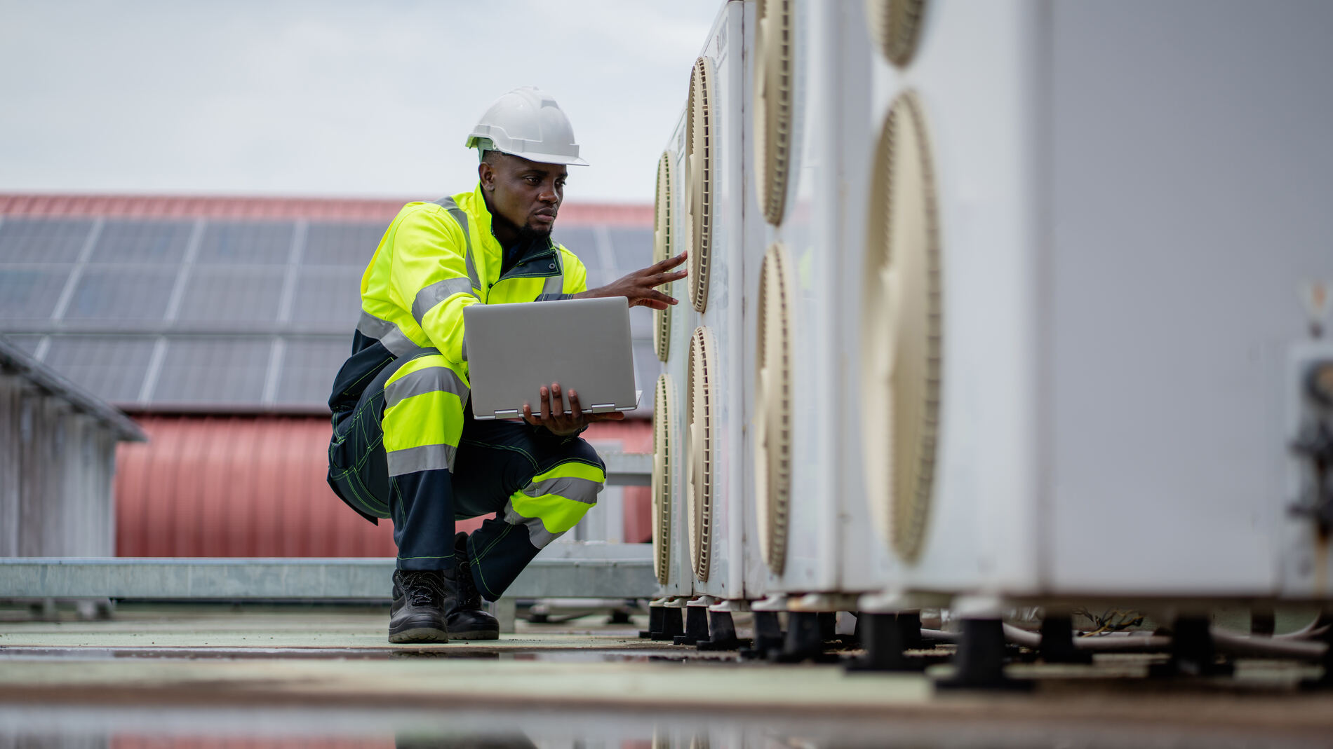 A technician wearing a white hard hat and yellow reflective safety jacket kneels while inspecting outdoor HVAC units, holding a laptop.