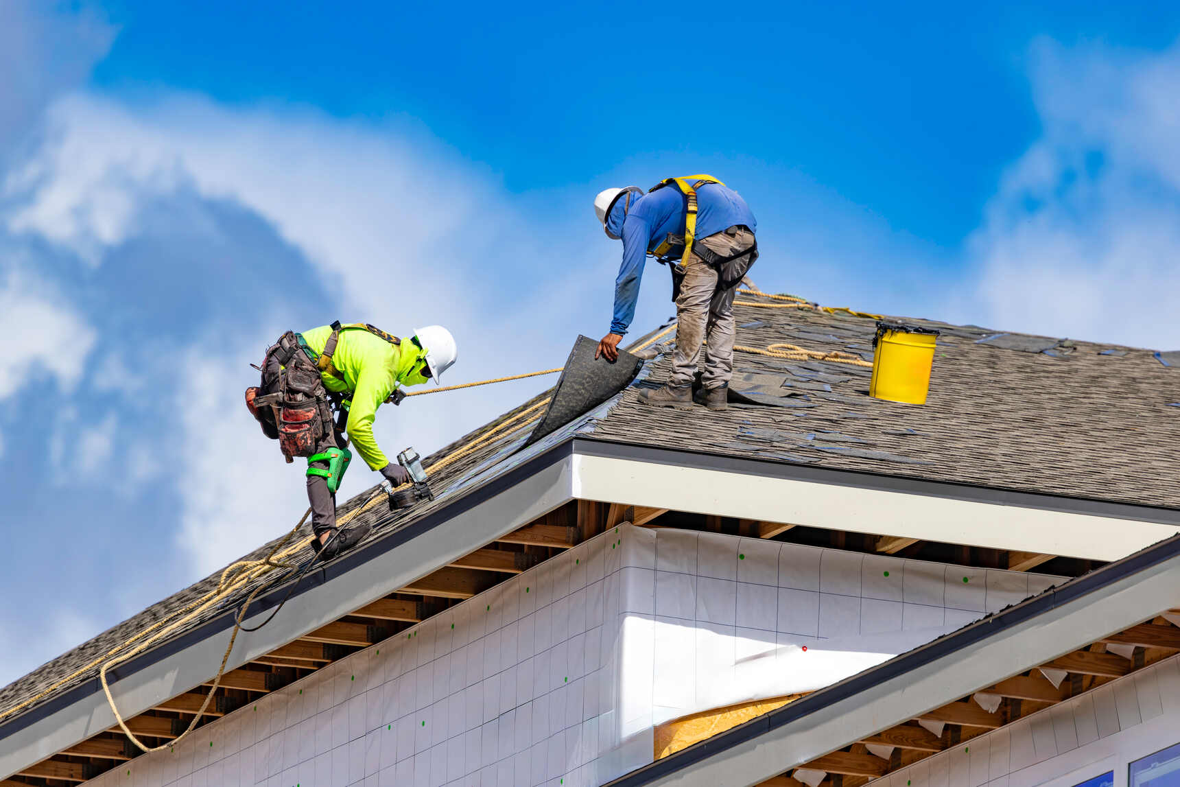 Two construction workers wearing safety harnesses and helmets install roofing materials on a house under clear blue sky, with one worker holding a rolled shingle and the other using a nail gun.